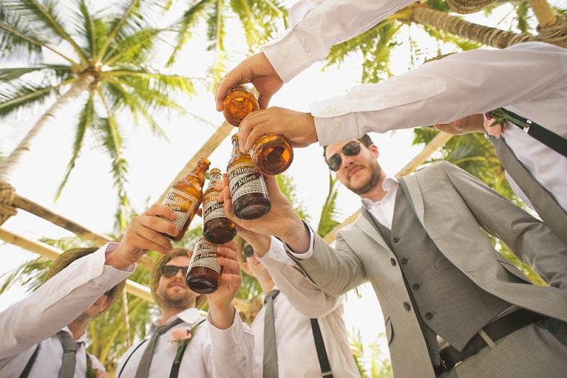 Male friends attending a beach wedding all holding beers performing a toast, view from bottom up