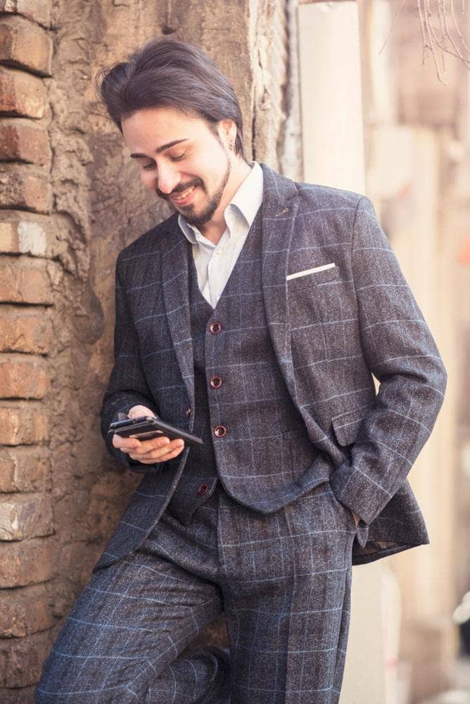 young fashionable man wearing tweed suit leaning against a wall using mobile phone